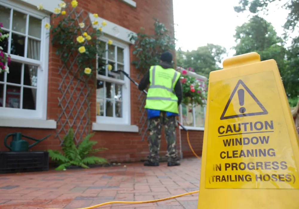 Window cleaner using a water-fed pole on a house with hanging baskets, beside a caution sign